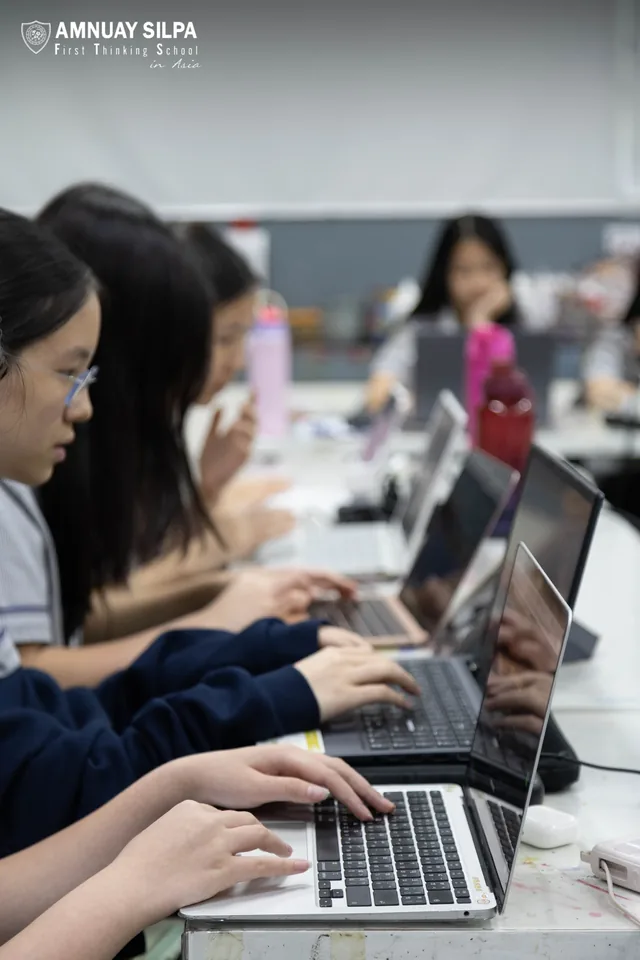 Students working on laptops at a classroom table with the Amnuay Silpa school logo visible at the top